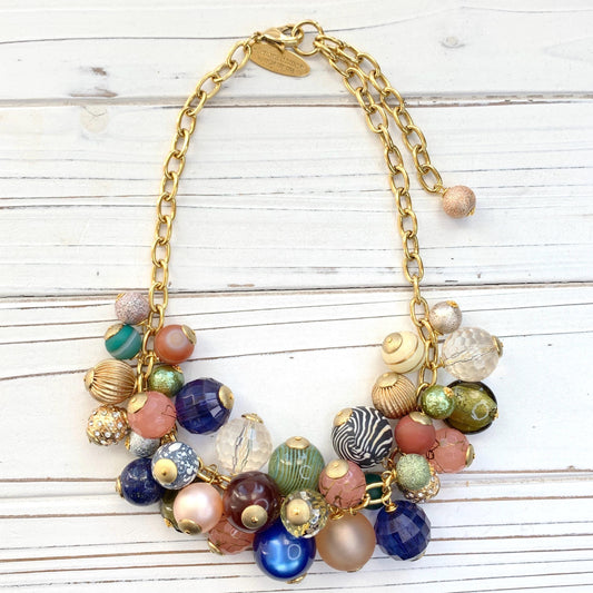 May include: Gold chain necklace featuring a cluster of colorful beads. Beads include various textures and patterns: clear, frosted, glittery, striped, and solid colors such as peach, teal, green, blue, brown, and cream. The necklace has a gold clasp. Lenora Dame necklace.