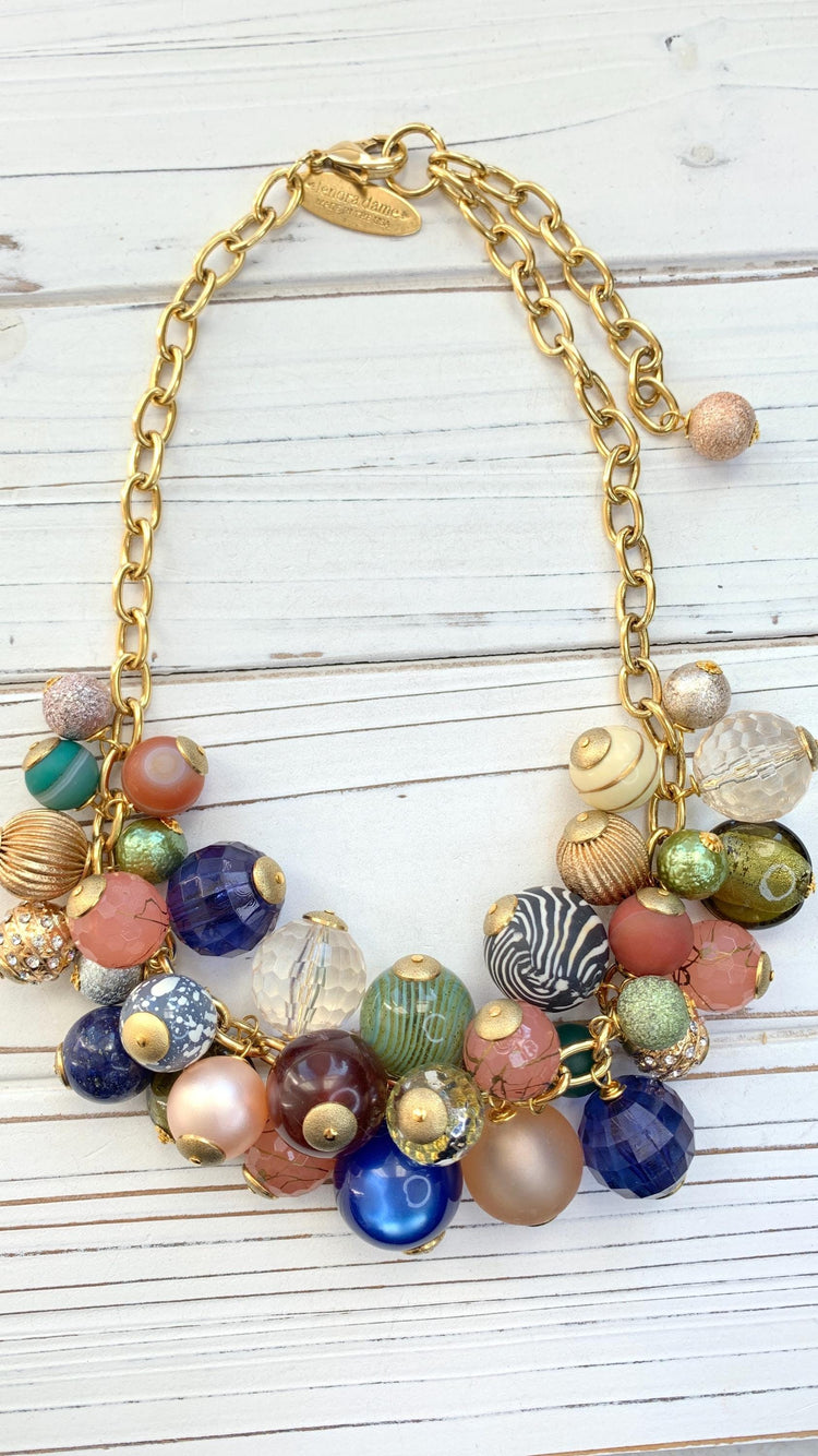 Gold chain necklace with multicolored beads. The beads are various shapes and sizes, including round, faceted, and textured. Colors include pink, peach, green, blue, brown, and clear. Some beads have gold accents. The necklace has a gold clasp with a small tag. The beads appear to be glass or resin.
