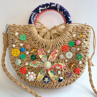 Small half-moon straw bag embellished with buttons, vintage earrings, and large metal flower broach decoupaged in red and yellow floral paper. Cute straw bag for summer with top handle and removable crossbody strap.