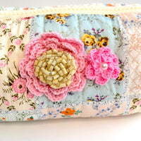 Floral cotton quilted makeup bag for women detailed with two pink crochet flowers, one with a beige vintage earring center and one with a pearl center. Small quilted makeup pouch with patchwork of floral prints, zipper and faux leather tassel zipper pull.
