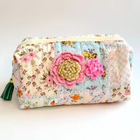 Floral cotton quilted makeup bag for women detailed with two pink crochet flowers, one with a beige vintage earring center and one with a pearl center. Small quilted makeup pouch with patchwork of floral prints, zipper and faux leather tassel zipper pull.