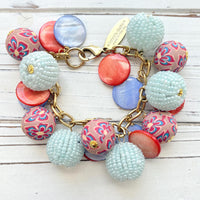 Chunky charm bracelet with assorted mix of light blue seed bead balls, floral fimo beads, blue and orange mother of pearl disc bead hung on a gold bracelet chain with lobster clasp and hangtag that says Lenora Dame Made in the USA. Lenora Dame jewelry made in USA.
