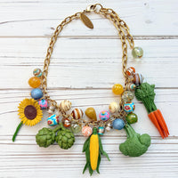 Colorful vegetable necklace for women with colorful beads and vegetable charms that include corn, artichokes, broccoli, carrots, and a sunflower hung on a gold chain along with a mix of colorful round beads. Gold chain included a hang tag that says Lenora Dame, Handmade in USA. Wacky necklace for women with vegetable charms shown on a white washed board. Lenora Dame necklace made in the USA.