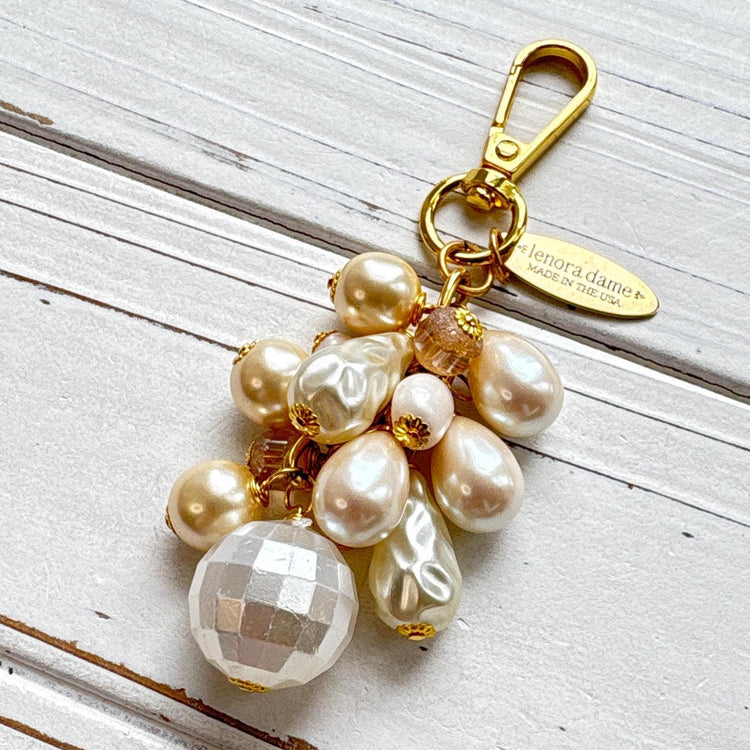 Pearl bag charm shown on a whitewashed board. A mix of cream and white faux pearls in different shapes are wire wrapped to a gold chain with gold clasp and oval hang tag that says Lenora Dame Made in USA. Lenora Dame bag charm.
