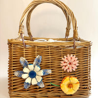 Top handle rectangular woven wicker handbag embellished with three metal flower brooches. One flower is blue and white, one pink flower brooch and one yellow and orange flower brooch. White fabric inner lining. Cute handbags for summer.