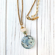 May include: A gold chain necklace with a round glass pendant featuring a blue and white pattern of flying birds. The pendant is attached to a white bead and a cluster of smaller blue beads. The necklace has a gold clasp and a small gold tag that reads 'lenora dame'.