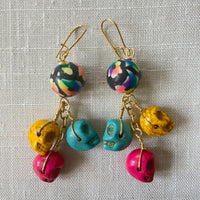 Lenora Dame sugar skull earrings with three miniature carved skulls in bright yellow, hot pink and turquoise wire wrapped under a black with rainbow flower design round polymer bead dangling from a gold French ear wire.