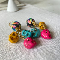 Lenora Dame sugar skull earrings with three miniature carved skulls in bright yellow, hot pink and turquoise wire wrapped under a black with rainbow flower design round polymer bead dangling from a gold French ear wire. 