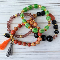 Lenora Dame 4-Piece Halloween beaded stretch bracelet set. Top set has oblong green glass beads with gold spacers. Second bracelet has orange beads with gold spacers, an orange thread tassel and a black and white thread tassel and a black tear drop bead adornment dangling from the left. The third bracelet has purple faceted beads. The last bracelet has a mix of black bumpy beads, purple faceted beads, larger round black beads and an orange faceted semiprecious stone.