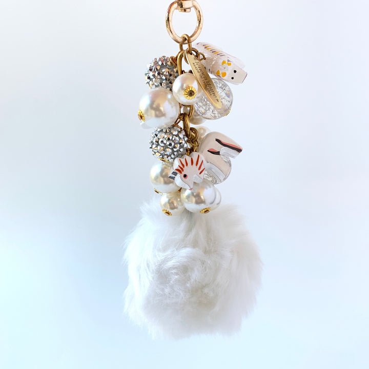 A white fluffy pom pom keychain with a gold chain and a variety of white, gold, and silver beads and charms. Lenora Dame bag charms.