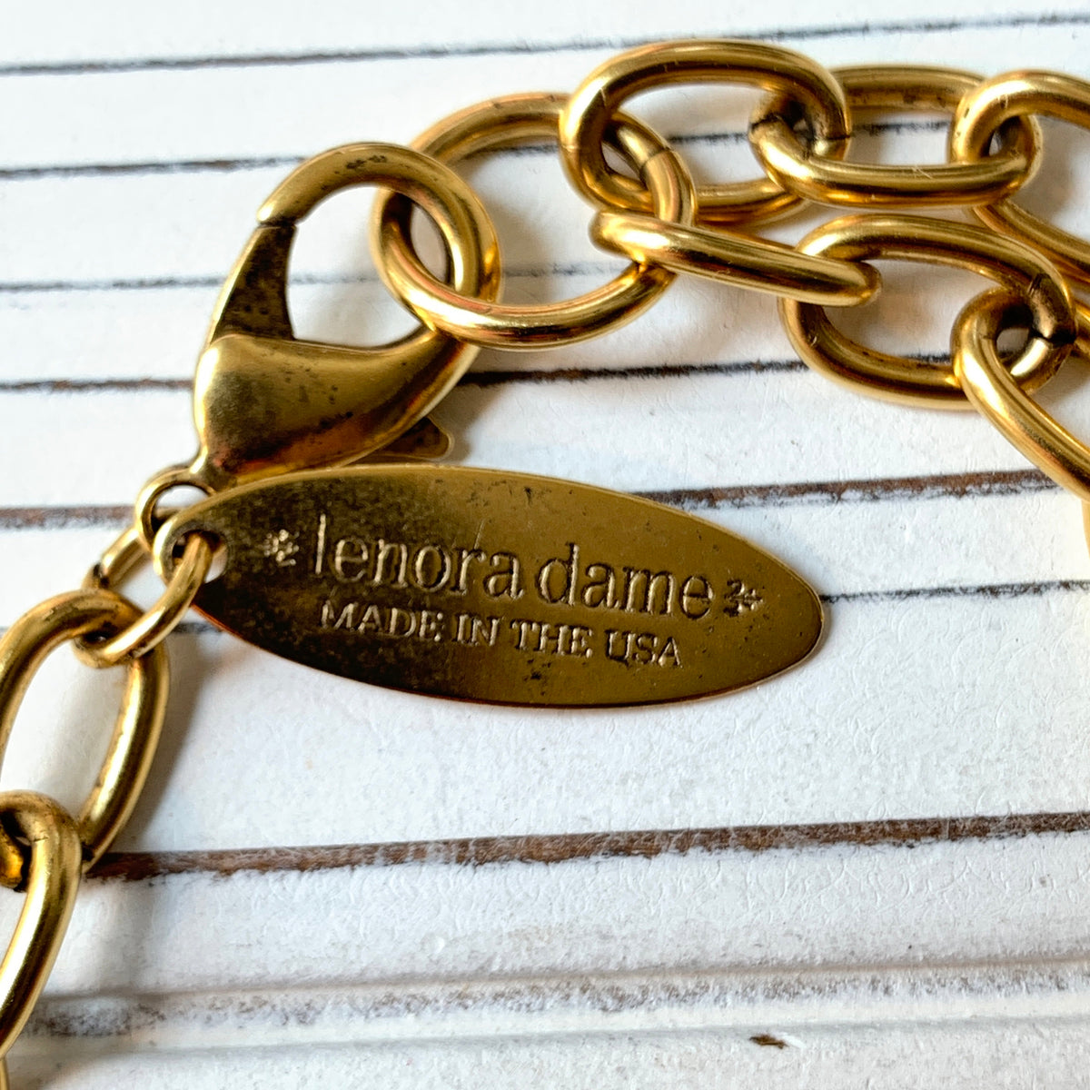 May include: A gold metal chain with a clasp and a tag that reads 'lenora dame* made in the usa'.