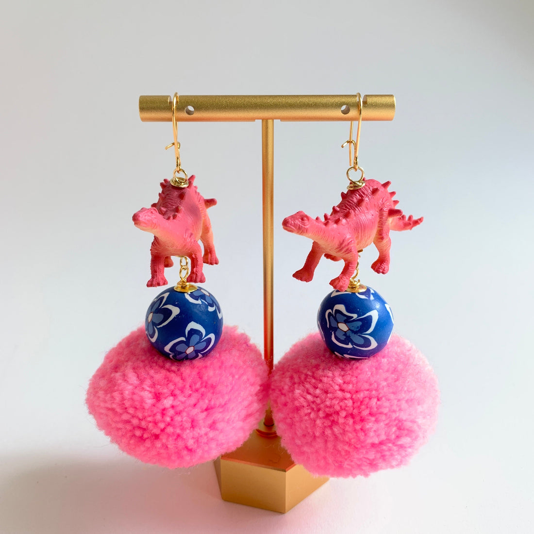Pink pom pom earrings with a blue and white floral ball and a pink dinosaur charm hanging from each earring. Lenora Dame dinosaur jewelry handmade in the USA.