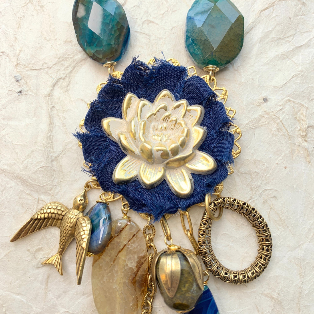 Lenora Dame necklace with metal white lotus mounted on navy blue fabric rosette and a mix of dangling semiprecious stones, brass bird charm and tear drop filigree piece.