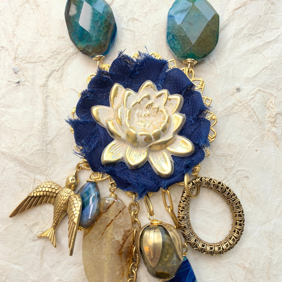 Lenora Dame necklace with metal white lotus mounted on navy blue fabric rosette and a mix of dangling semiprecious stones, brass bird charm and tear drop filigree piece.