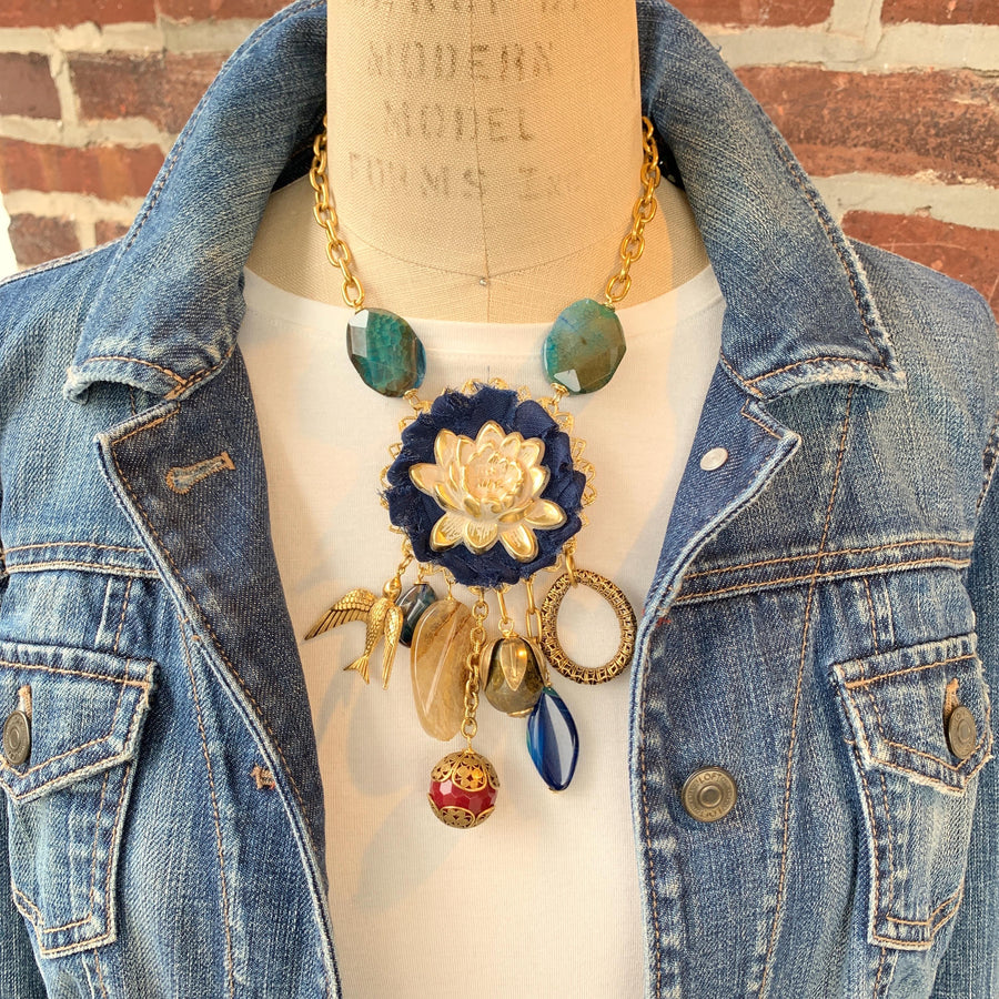 Lenora Dame necklace with metal white lotus mounted on navy blue fabric rosette and a mix of dangling semiprecious stones, brass bird charm and tear drop filigree piece.