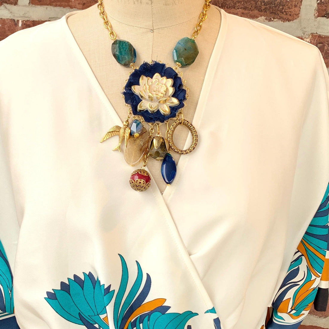 Lenora Dame necklace with metal white lotus mounted on navy blue fabric rosette and a mix of dangling semiprecious stones, brass bird charm and tear drop filigree piece.