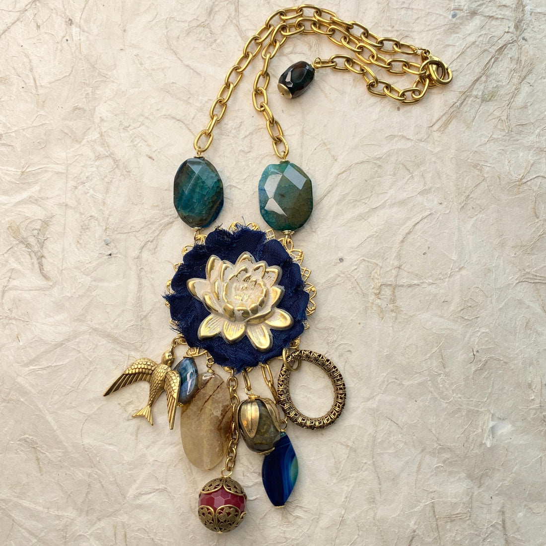 Lenora Dame necklace with metal white lotus mounted on navy blue fabric rosette and a mix of dangling semiprecious stones, brass bird charm and tear drop filigree piece.