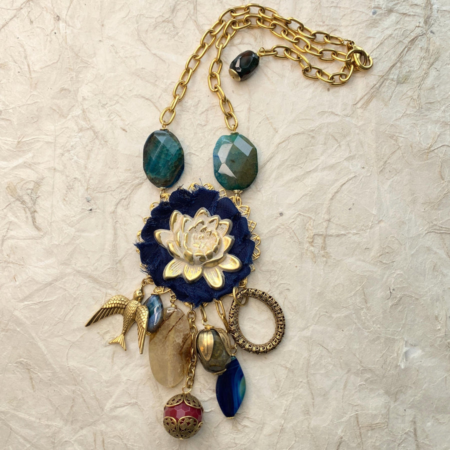 Lenora Dame necklace with metal white lotus mounted on navy blue fabric rosette and a mix of dangling semiprecious stones, brass bird charm and tear drop filigree piece.