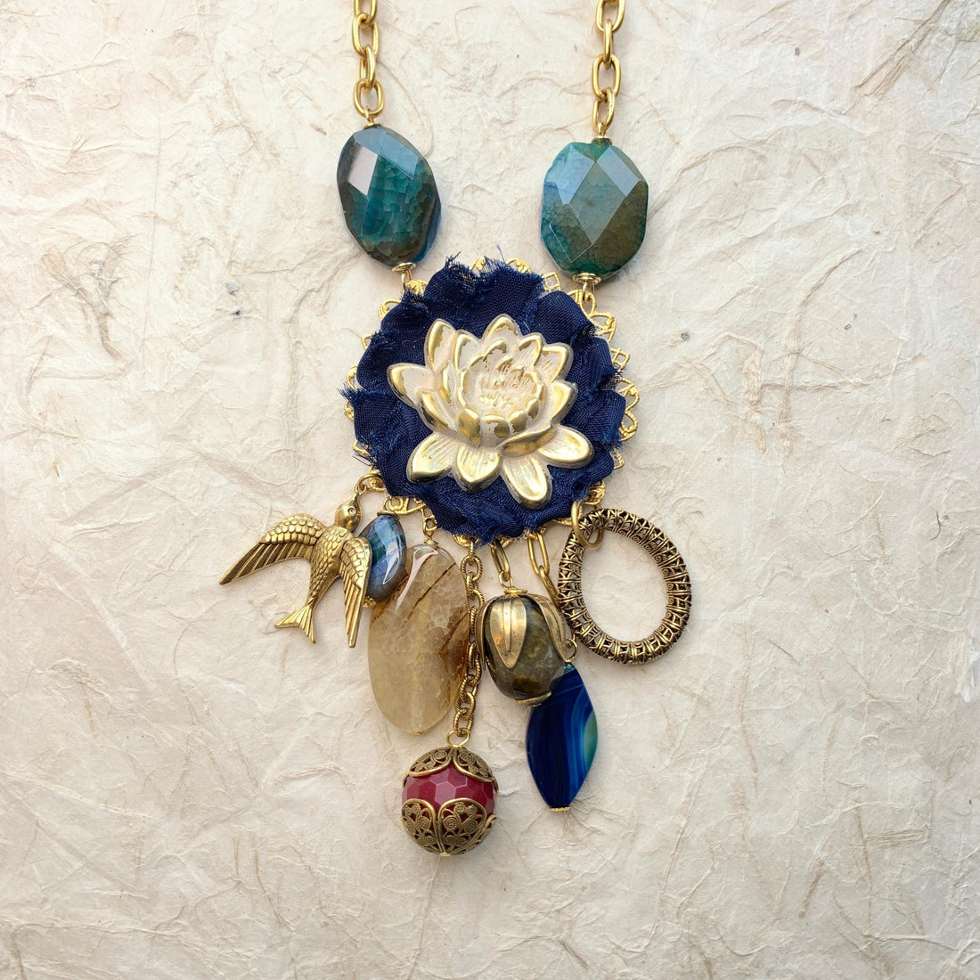 Lenora Dame necklace with metal white lotus mounted on navy blue fabric rosette and a mix of dangling semiprecious stones, brass bird charm and tear drop filigree piece.