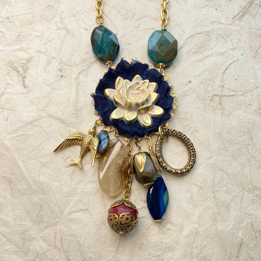 Lenora Dame necklace with metal white lotus mounted on navy blue fabric rosette and a mix of dangling semiprecious stones, brass bird charm and tear drop filigree piece.