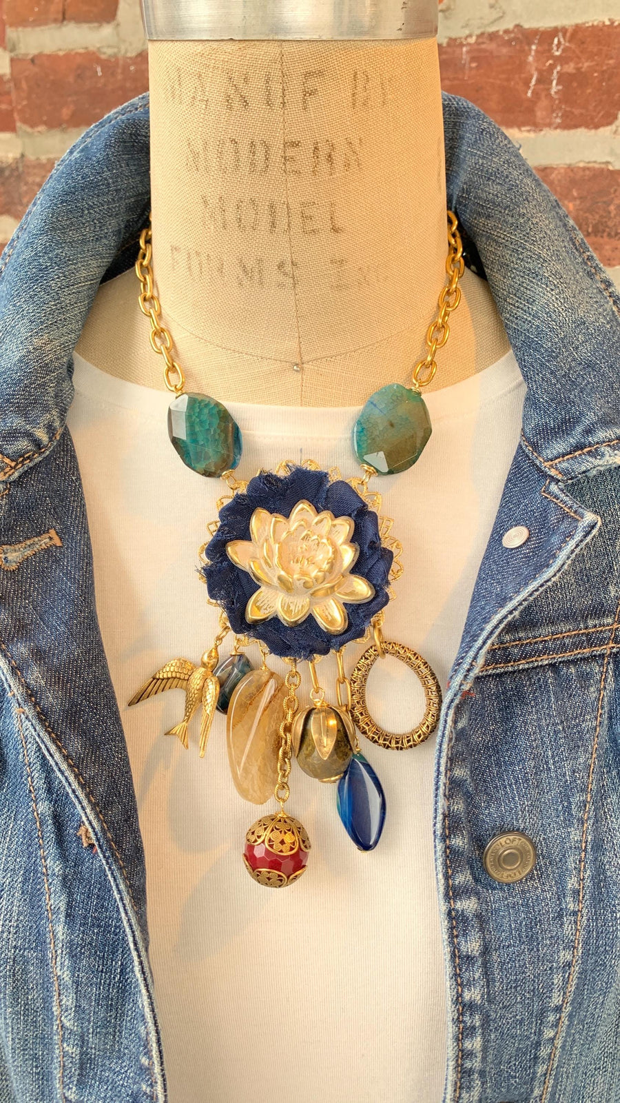 Lenora Dame necklace with metal white lotus mounted on navy blue fabric rosette and a mix of dangling semiprecious stones, brass bird charm and tear drop filigree piece.