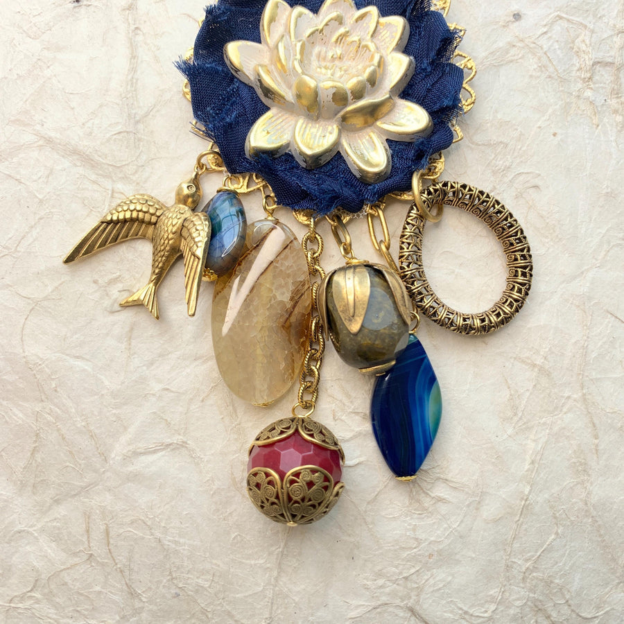 Lenora Dame necklace with metal white lotus mounted on navy blue fabric rosette and a mix of dangling semiprecious stones, brass bird charm and tear drop filigree piece.