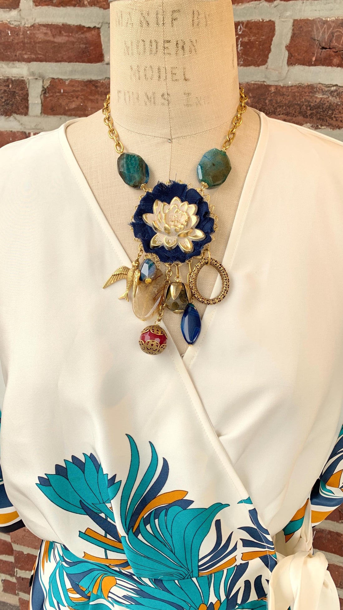 Lenora Dame necklace with metal white lotus mounted on navy blue fabric rosette and a mix of dangling semiprecious stones, brass bird charm and tear drop filigree piece.