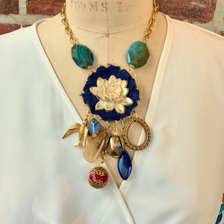 White lotus necklace. One of a kind handmade necklace by Lenora Dame. Lenora Dame necklace with metal white lotus mounted on navy blue fabric rosette and a mix of dangling semiprecious stones, brass bird charm and tear drop filigree piece.