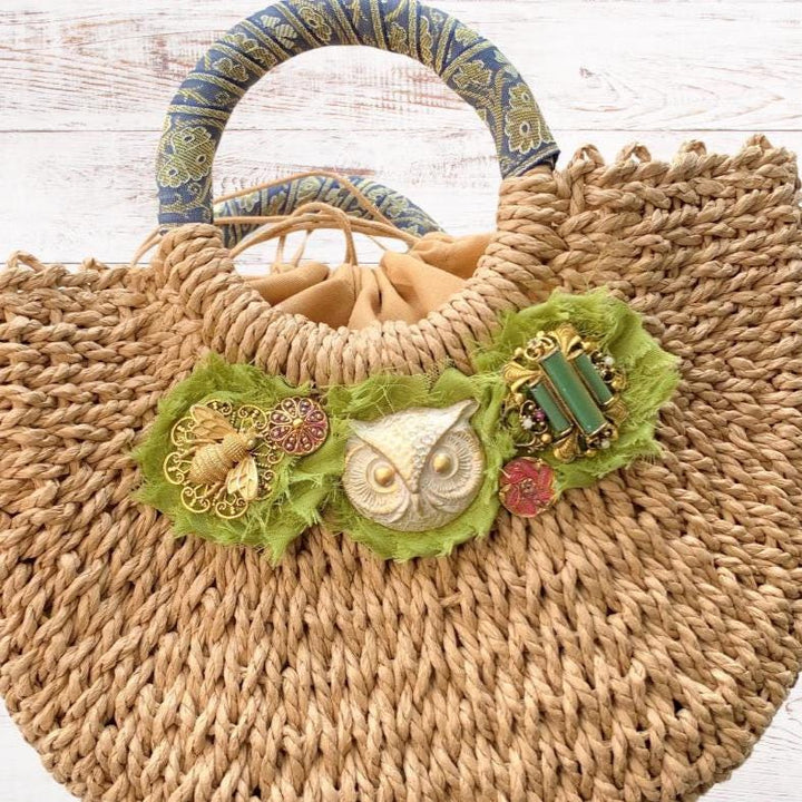 Woven straw handbag with contrast top handle, three green frayed edge rosettes topped with a gold bee, white painted gold metal owl stamping and a vintage brooch. Cute straw bags for summer. 