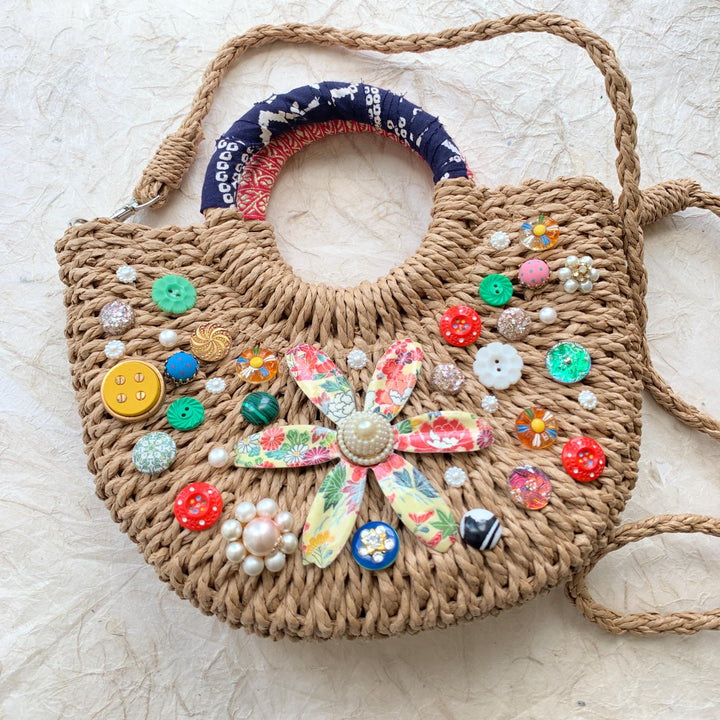 Small half-moon straw bag embellished with buttons, vintage earrings, and large metal flower broach decoupaged in red and yellow floral paper. Cute straw bag for summer with top handle and removable crossbody strap.