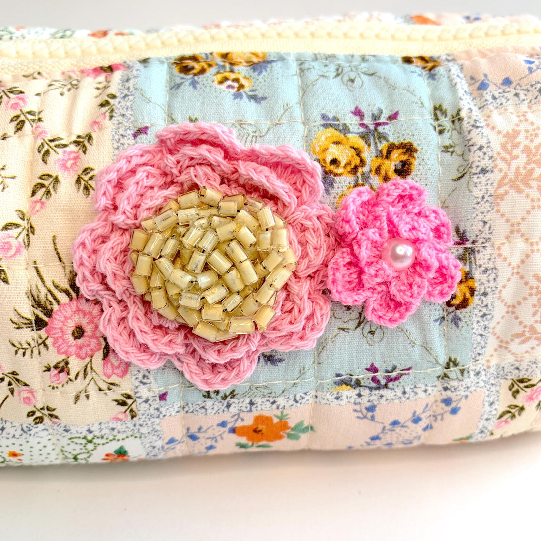 Floral cotton quilted makeup bag for women detailed with two pink crochet flowers, one with a beige vintage earring center and one with a pearl center. Small quilted makeup pouch with patchwork of floral prints, zipper and faux leather tassel zipper pull.