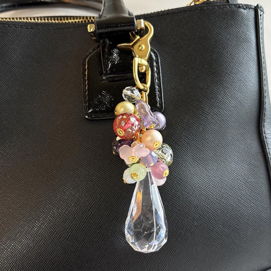 Pastel beaded bag charm with gold trigger clasp, a mix of beads and small glass birds above a large clear faceted teardrop shaped Lucite crystal shown hung on a black handbag. Lenora Dame bag charm.