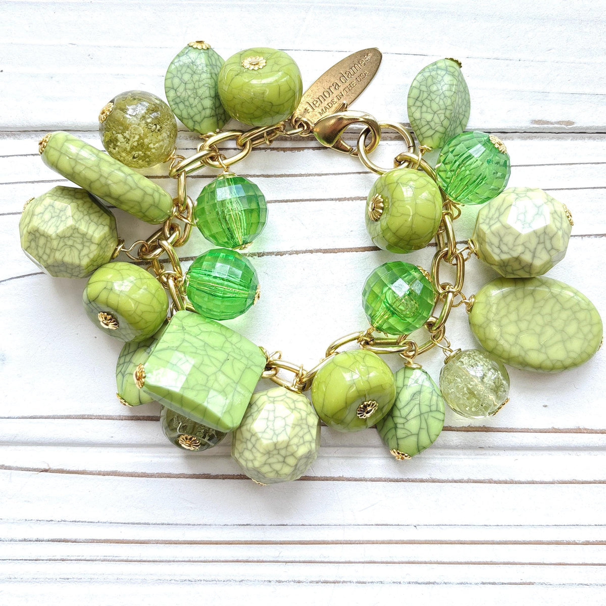 Gold chain charm bracelet filled with assorted green beads, some beads have a crackle look to them. Bracelet has a hangtag that says Lenora Dame, Handmade in the USA.