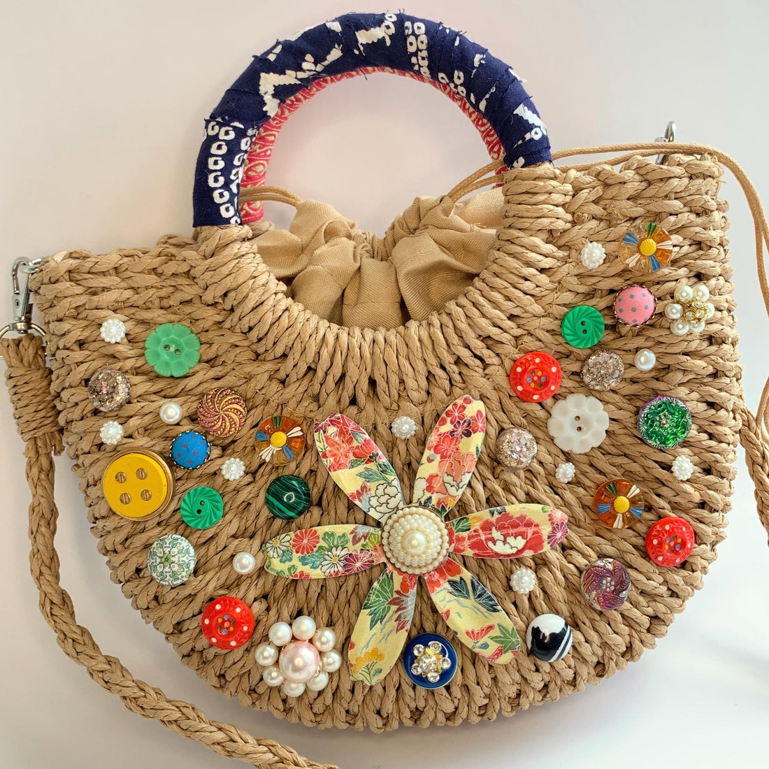 Small half-moon straw bag embellished with buttons, vintage earrings, and large metal flower broach decoupaged in red and yellow floral paper. Cute straw bag for summer with top handle and removable crossbody strap.