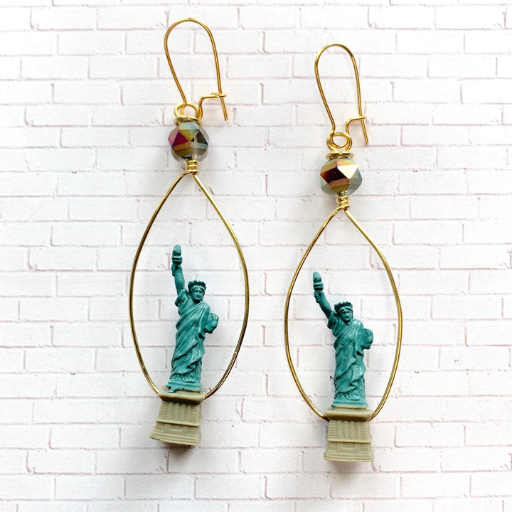 Statue of liberty earrings by Lenora Dame.  A pair of gold hoop earrings with a teal blue Statue of Liberty charm hanging from each hoop. Each charm has a gold base and a clear iridescent bead at the top of the hoop.