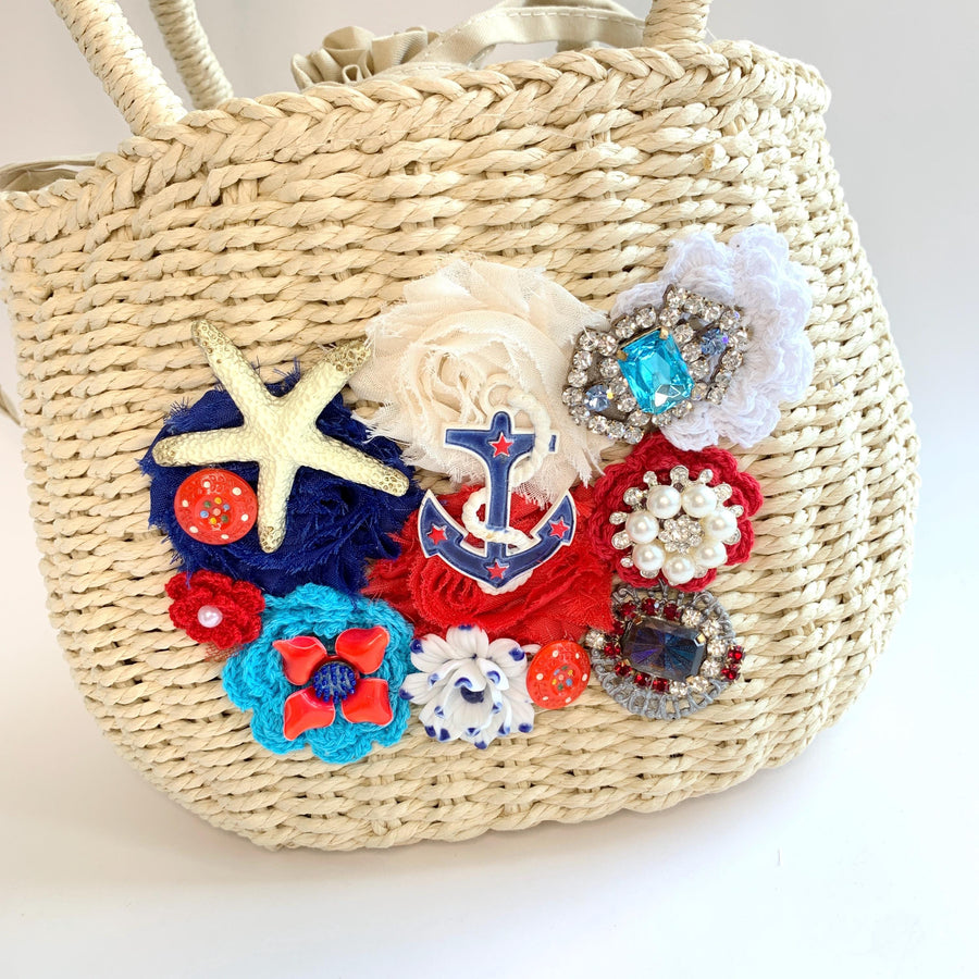 Nautical tote bag detailed with red, white and blue frayed edge fabric rosettes, star fish, anchor, blue rhinestone, and vintage buttons. Light beige woven handbag with drawstring fabric inner lining and top handle. Straw summer handbag.