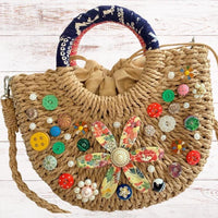 Small half-moon straw bag embellished with buttons, vintage earrings, and large metal flower broach decoupaged in red and yellow floral paper. Cute straw bag for summer with top handle and removable crossbody strap.