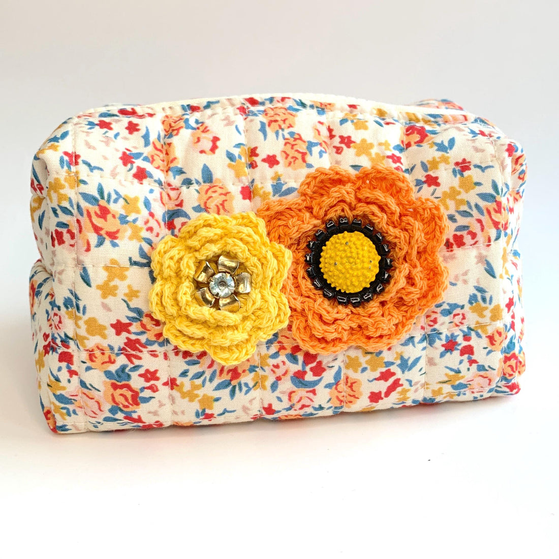 Floral cotton quilted makeup bag for women detailed with yellow and orange crochet flowers with button and rhinestone centers. Small quilted makeup pouch with orange, blue and red floral print.