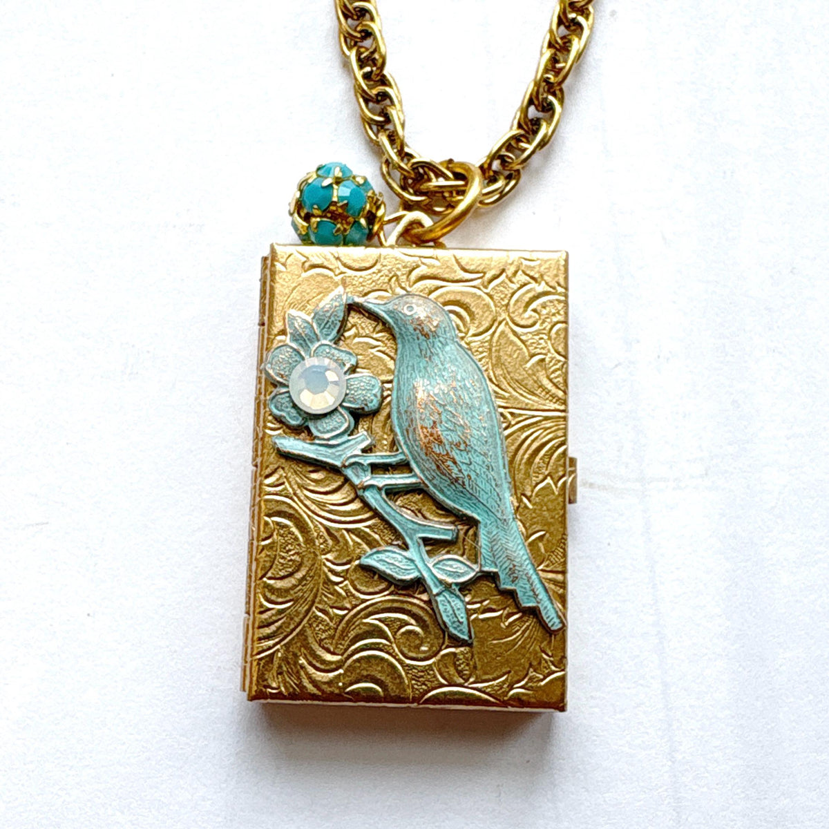 Brocade etched gold photo locket necklace with a gold rectangle locket mounted with a metal bird on a flower stem design painted an green aqua color with detail on the flower hung on a gold chain with a  opaque turquoise rhinestone bead ball. Gold locket for women. Lenora Dame necklace made in the USA.