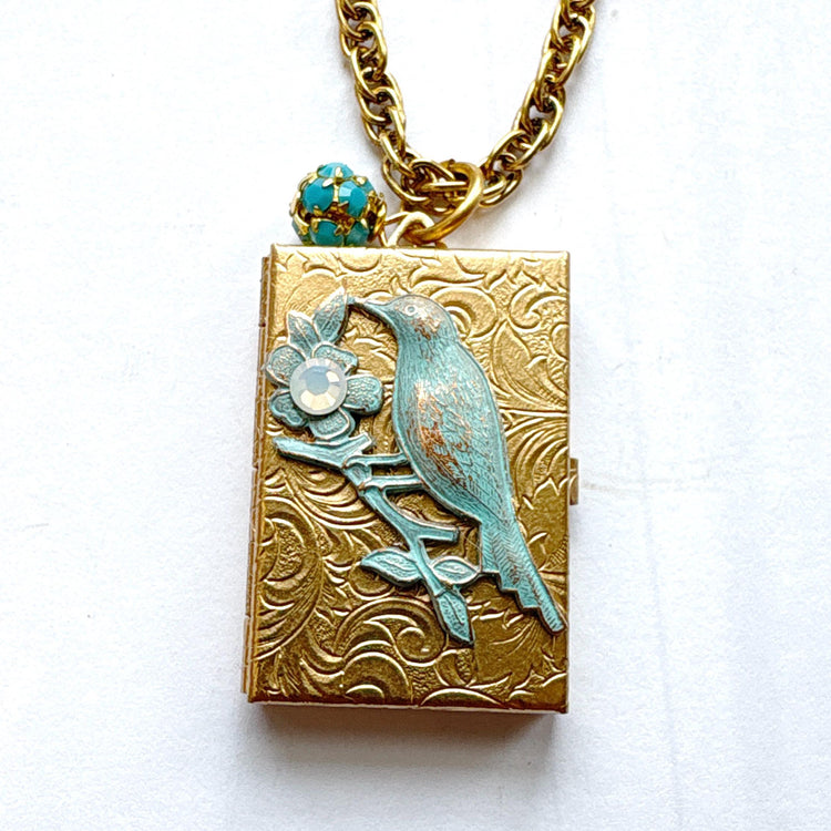 Brocade etched gold photo locket necklace with a gold rectangle locket mounted with a metal bird on a flower stem design painted an green aqua color with detail on the flower hung on a gold chain with a  opaque turquoise rhinestone bead ball. Gold locket for women. Lenora Dame necklace made in the USA.