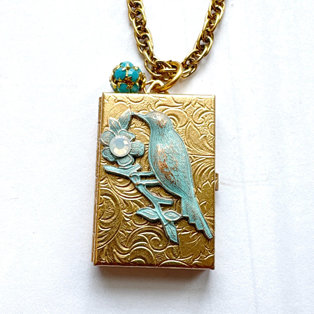 Brocade etched gold photo locket necklace with a gold rectangle locket mounted with a metal bird on a flower stem design painted an green aqua color with detail on the flower hung on a gold chain with a  opaque turquoise rhinestone bead ball. Gold locket for women. Lenora Dame necklace made in the USA.
