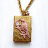 Brocade etched gold photo locket necklace with a gold rectangle locket mounted with a metal bird on a flower stem design painted pink with rhinestone detail on the flower hung on a gold chain with a pink rhinestone bead ball. Gold locket for women. Lenora Dame necklace made in the USA.