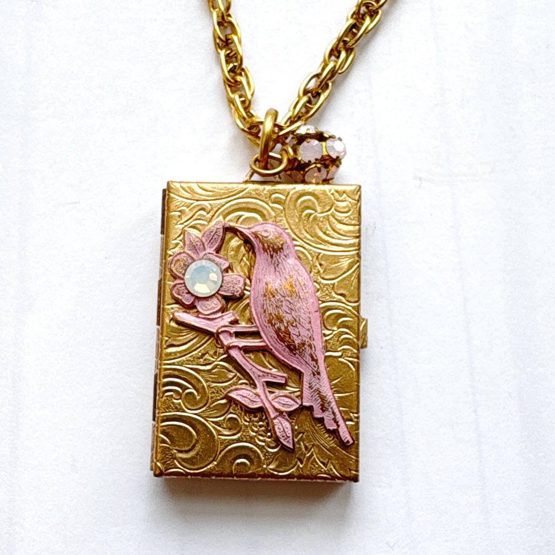Brocade etched gold photo locket necklace with a gold rectangle locket mounted with a metal bird on a flower stem design painted pink with rhinestone detail on the flower hung on a gold chain with a pink rhinestone bead ball. Gold locket for women. Lenora Dame necklace made in the USA.