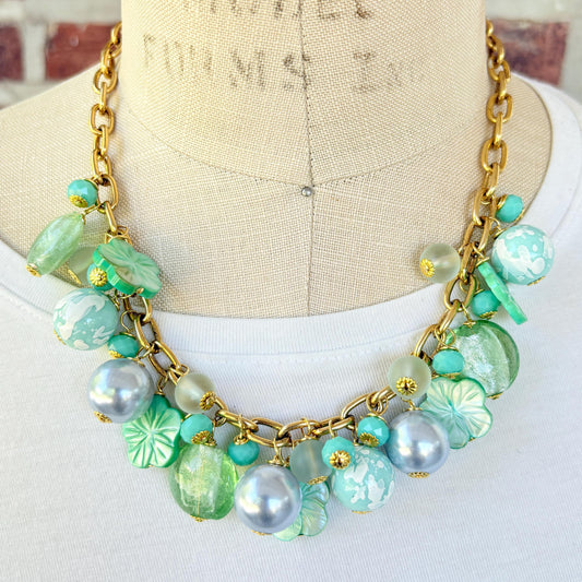 A mix of light green and light blue beads, pearls, and mother of pearl flowers hand from a gold chain necklace shown on a dress form wearing a white shirt. Green beaded necklace for women made in the USA. Lenora Dame necklace.