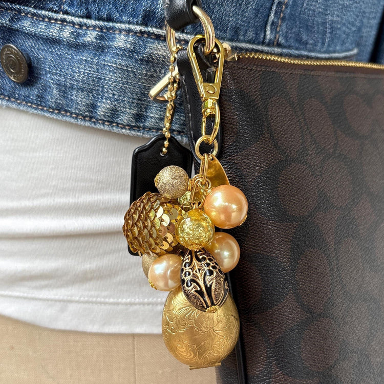 Gold tone beaded bag charm with a etched brass egg shaped hinged photo locket below a mix of pearls, lampworked glass beads, a gold sequin beads, and a black bead with gold scroll design. The beads hang on a gold chain with a gold clasp and Lenora Dame Made in USA hang tag. Lenora Dame bag charm. Bag charm is shown hung on a brown crossbody bag.
