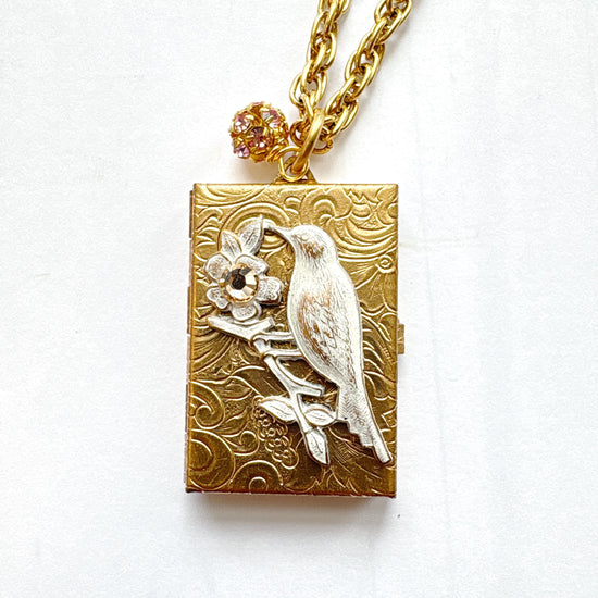 Brocade etched gold photo locket necklace with a gold rectangle locket mounted with a metal bird on a flower stem design painted white with rhinestone detail on the flower hung on a gold chain with a rhinestone bead ball. Gold locket for women. Lenora Dame necklace made in the USA.