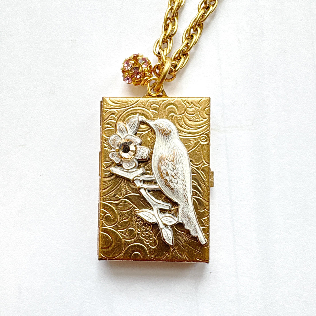 Brocade etched gold photo locket necklace with a gold rectangle locket mounted with a metal bird on a flower stem design painted white with rhinestone detail on the flower hung on a gold chain with a rhinestone bead ball. Gold locket for women. Lenora Dame necklace made in the USA.