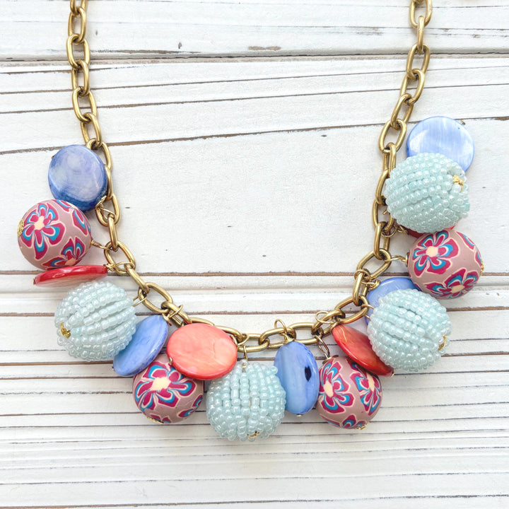 Chunky charm necklace with assorted mix of light blue seed bead balls, floral fimo beads, blue and orange mother of pearl disc bead hung on a gold necklace chain with lobster clasp and hangtag that says Lenora Dame Made in the USA. Lenora Dame necklace.
