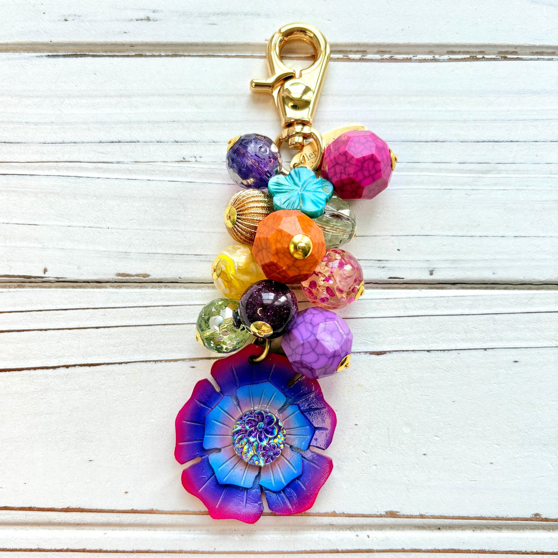Colorful beaded bag charm filled with assorted beads in different shapes in vibrant colors. A large pink, purple and blue flower is at the bottom of the bead cluster. A gold trigger clasp is above the beads. Lenora Dame bag charms.