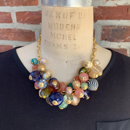 May include: Statement necklace featuring a gold chain and an array of multicolored beads. The beads are varied in size, shape, and pattern, including opaque and translucent textures. Colors include teal, pink, gold, brown, and dark blue.  The necklace is displayed on a dress form.